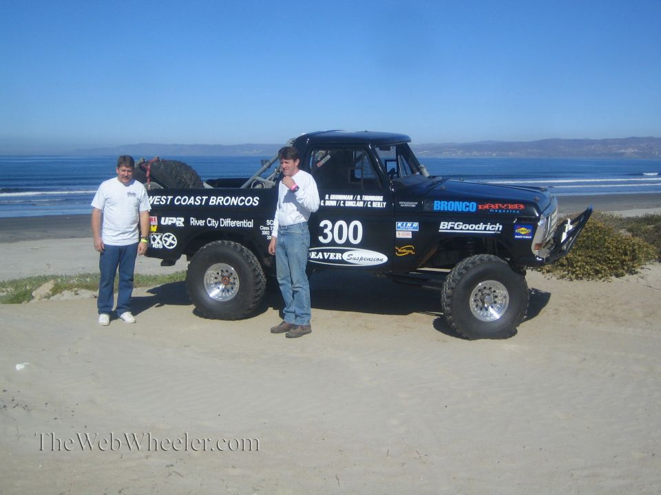 Two men stand beside a black number 300 West Coast Broncos race truck on a beach