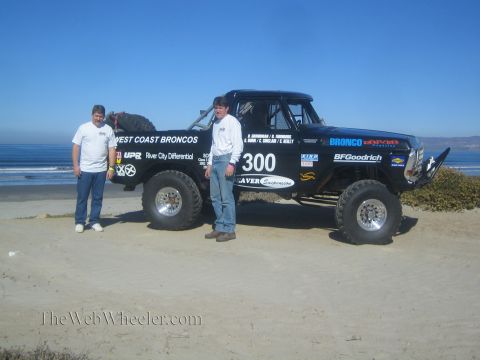 1990 Ford Bronco - Two men in white shirts stand at the side of black race truck 300 with West Coast Broncos livery on a beach by the ocean.