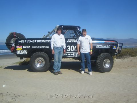 1990 Ford Bronco - Two men in white shirts and jeans stand in front of the black number 300 West Coast Broncos off-road race truck on the sand.