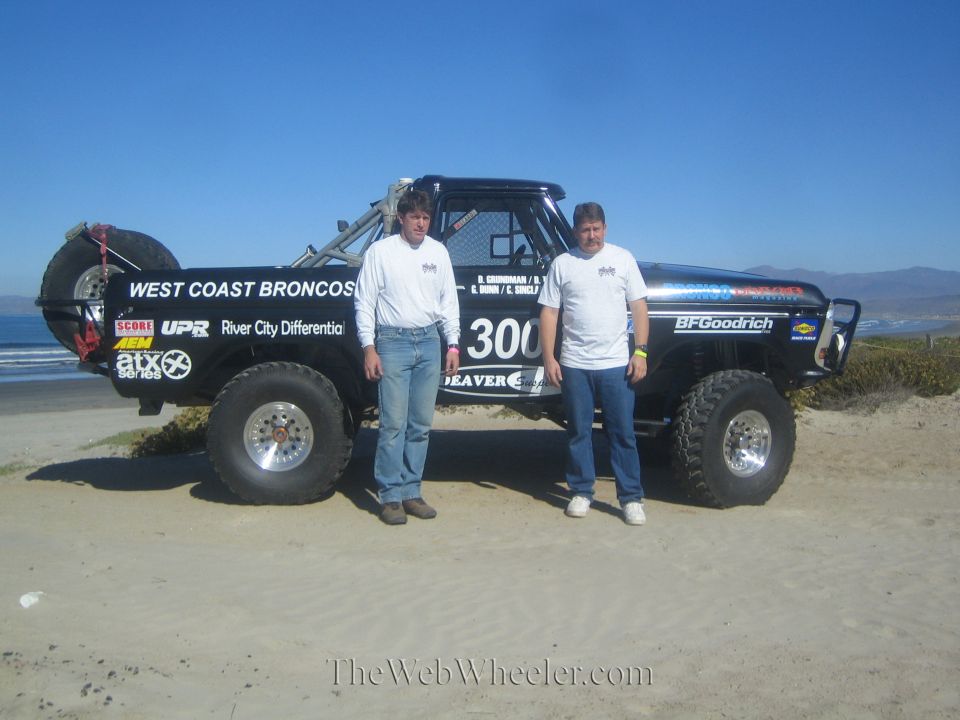 Two men pose in front of a black number 300 West Coast Broncos race truck on the beach