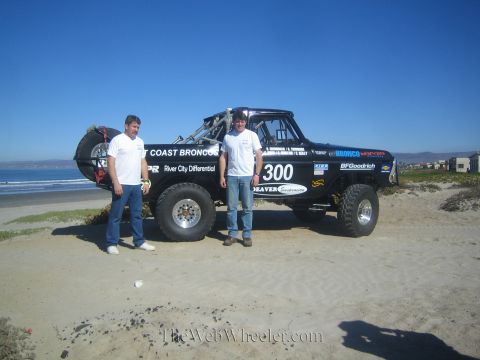 1990 Ford Bronco - Two men pose in front of the black West Coast Broncos number 300 race truck parked on a beach with ocean and hills behind.