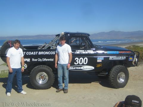 1990 Ford Bronco - Two men stand by the black number 300 race truck on the beach while a camera lens appears at lower right framing the shot.