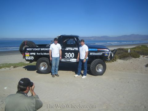1990 Ford Bronco - crouching photographer shoots two men posing beside the black number 300 West Coast Broncos race truck on a coastal beach.