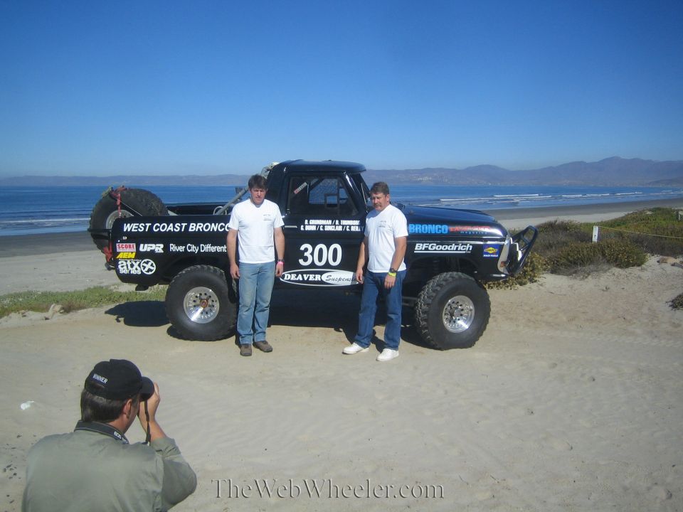 Two men pose with black number 300 race truck as a photographer crouches in the foreground