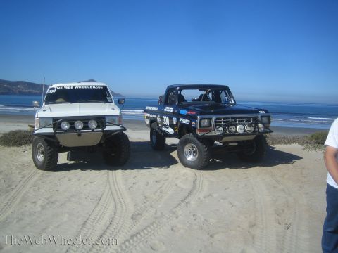 1990 Ford Bronco - white lifted SUV with a light bar sits beside the black number 300 off-road race truck on the sand near the ocean surf.