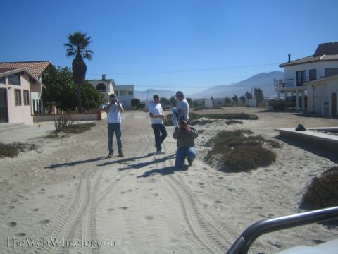 1990 Ford Bronco - Three people stand on a sandy road in a coastal village, one with a camera, with single-story houses, a palm tree and hills beyond.