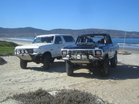 1990 Ford Bronco - white lifted Bronco-style SUV and a dark blue number 300 off-road truck sit side by side on a beach dune with the ocean behind.