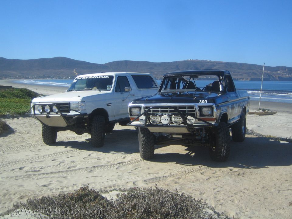 White lifted Bronco SUV and blue number 300 off-road truck parked together on a beach dune