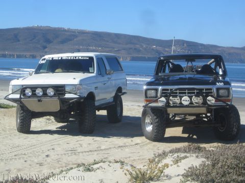 1990 Ford Bronco - white lifted SUV and a dark number 300 race truck, both with front light bars, sit on the sand with ocean and hills behind.