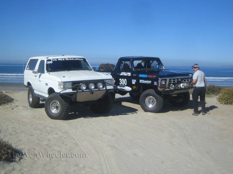1990 Ford Bronco - Two lifted Ford Broncos, one white and one black race-prepped with number 300, parked on sand by the ocean as a man stands nearby.