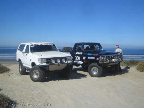 1990 Ford Bronco - white lifted Bronco and a black race-prepped Bronco numbered 300 sit on the sand near the shoreline with a person standing at right.