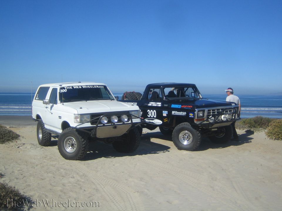 White and black lifted Ford Bronco trucks on a beach with ocean behind