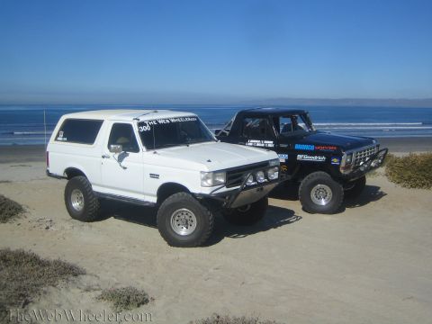 1990 Ford Bronco - white lifted Ford Bronco beside a black off-road Bronco with sponsor decals, parked on the beach with the ocean in the background.