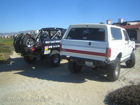 1990 Ford Bronco - Rear three-quarter view of a white lifted Ford Bronco numbered 300 beside a tube-chassis race truck with spare tires, on a dirt lot.