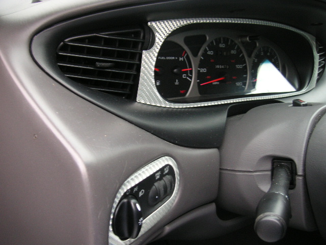 Dashboard interior of a 1999 Ford Taurus, showing the instrument cluster, air vent, and headlight controls with carbon fiber trim