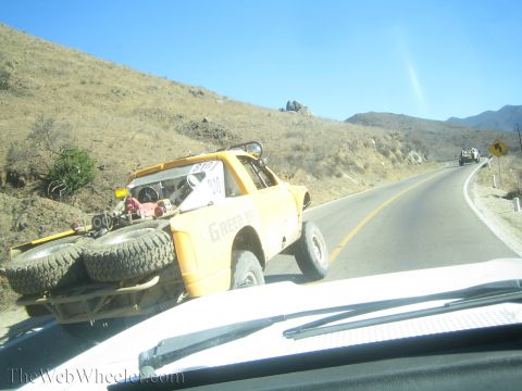 1990 Ford Bronco - yellow off-road race truck loaded with spare tires drives along a winding paved mountain road, seen through a windshield.