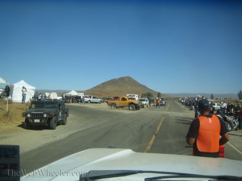 1990 Ford Bronco - roadside pit area in the desert with canopy tents, a Humvee, parked trucks, and spectators in safety vests near a small hill.