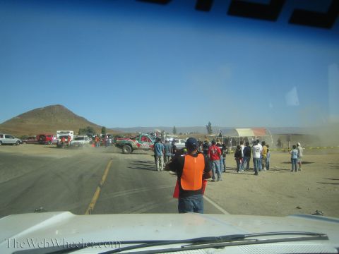 1990 Ford Bronco - Crowds of spectators and a marshal in an orange vest watch dust-kicking trucks at a desert roadside pit area below a hill.