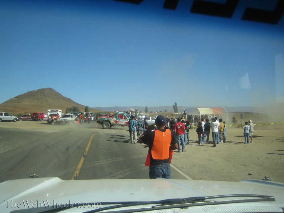 Spectators in orange vests watching dusty trucks at a desert pit stop