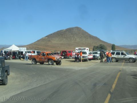 1990 Ford Bronco - orange off-road pickup and various support vehicles and tents gather at a dusty desert intersection beneath a conical hill.