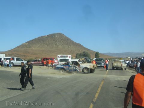1990 Ford Bronco - mud-coated race truck slides through dust at a desert pit area as marshals and spectators stand near an ambulance and parked trucks.