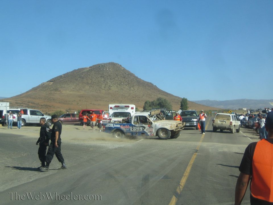 Mud-covered race truck sliding through a dusty desert pit with marshals