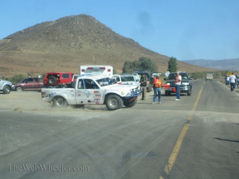 1990 Ford Bronco - white sponsor-covered race truck kicks up dust at a desert roadside pit, with an ambulance, support trucks, and marshals nearby.