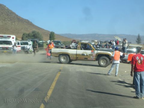 1990 Ford Bronco - tan-and-white race pickup throws up dust at a crowded desert pit area surrounded by marshals, spectators, and support vehicles.