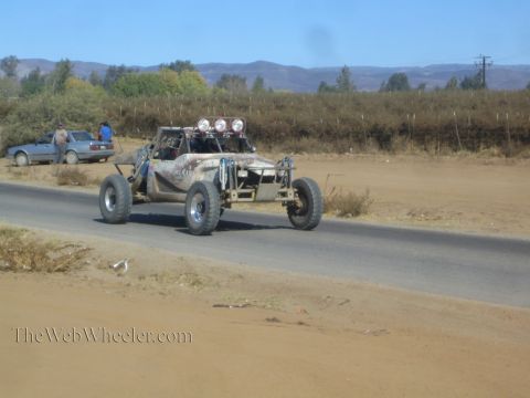 1990 Ford Bronco - weathered open-cockpit off-road race buggy with roof lights drives a paved road past a parked car and dry hills.
