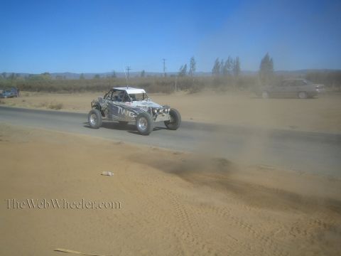 1990 Ford Bronco - off-road race buggy speeds along a paved road kicking up dust and sand, with a passing car visible in the background.