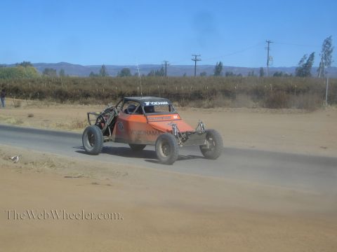 1990 Ford Bronco - orange single-seat off-road race buggy with Yokohama livery drives along a paved road beside a sandy dirt shoulder.