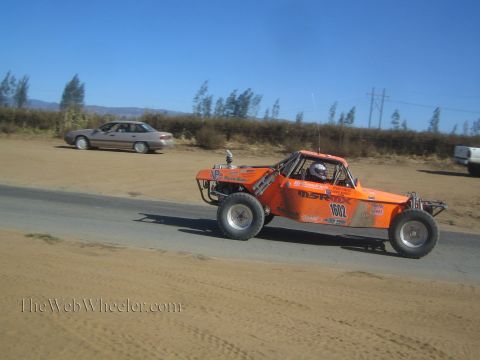 1990 Ford Bronco - orange off-road race buggy numbered 1602 with a helmeted driver travels a dirt-edged paved road past a parked silver car.
