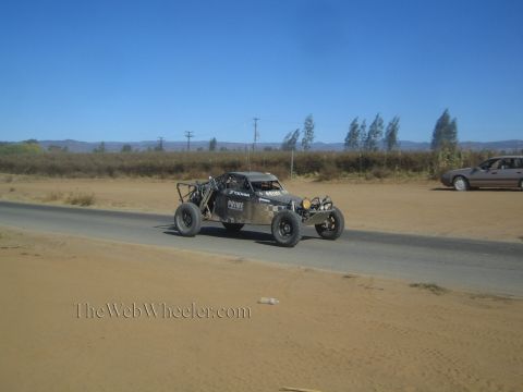 1990 Ford Bronco - dark off-road race buggy labeled Prime drives a sandy-shouldered paved road as a silver car passes in the background.