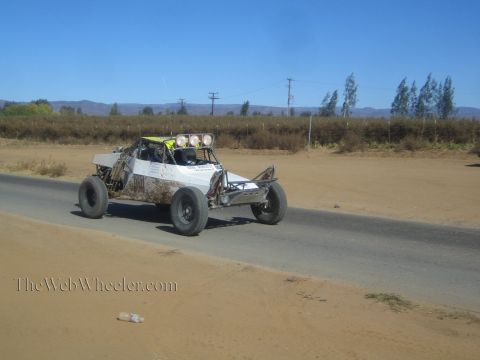 1990 Ford Bronco - white open-cockpit off-road race buggy with roof-mounted lights drives along a paved road bordered by sand and dry brush.
