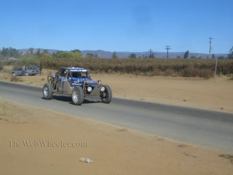 1990 Ford Bronco - blue-and-silver off-road race buggy with a helmeted driver speeds a paved road past a parked truck and dry fields.
