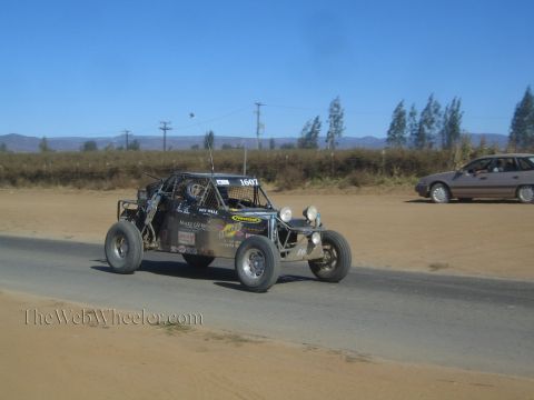 1990 Ford Bronco - dark off-road race buggy numbered 1607 with roof lights drives a paved road as a silver car passes on the dirt shoulder behind.