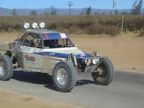 1990 Ford Bronco - blue-and-silver off-road race buggy numbered 1512 with Toto Motor Sports livery and roof lights drives a paved road by dry fields.