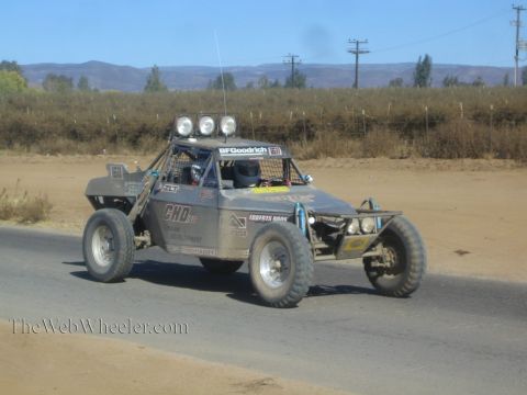1990 Ford Bronco - gray sponsor-decaled off-road race buggy with roof-mounted lights drives a paved road bordered by sandy shoulders and dry hills.