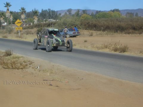 1990 Ford Bronco - dark off-road race buggy with Monster Energy graphics drives a paved road past a parked pickup, palms, and dry hills.