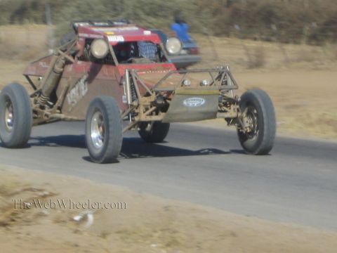 1990 Ford Bronco - red tube-frame off-road race buggy speeds along a paved desert road, its front wheels lifting off the pavement.