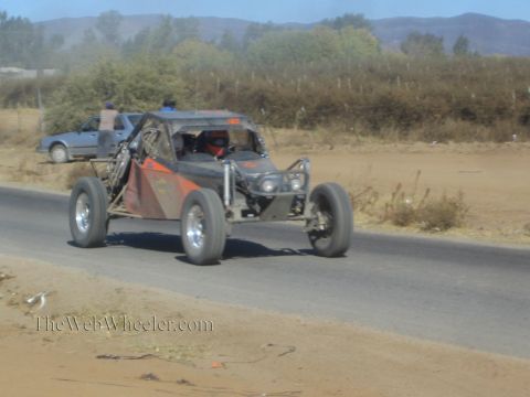 1990 Ford Bronco - black and orange single-seat off-road race buggy travels a paved road, with a sedan and onlookers in the background.