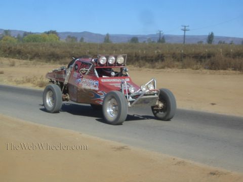 1990 Ford Bronco - red off-road race buggy with flame graphics and a roof light bar drives a paved road beside a dirt shoulder.