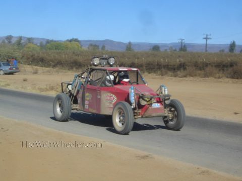 1990 Ford Bronco - weathered red off-road race buggy with twin roof lights races down a paved road in a dry rural landscape.