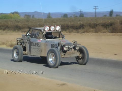 1990 Ford Bronco - gray off-road race buggy numbered 1644 with three roof lights speeds along a paved desert road.
