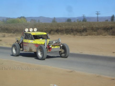 1990 Ford Bronco - yellow and red off-road race buggy with roof lights races along a paved road in dry hilly terrain.