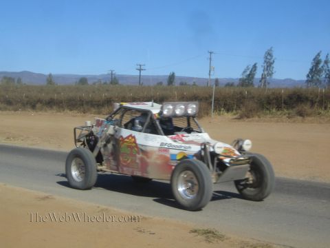 1990 Ford Bronco - white off-road race buggy with red graphics and three roof lights drives a paved road beside a dirt shoulder.