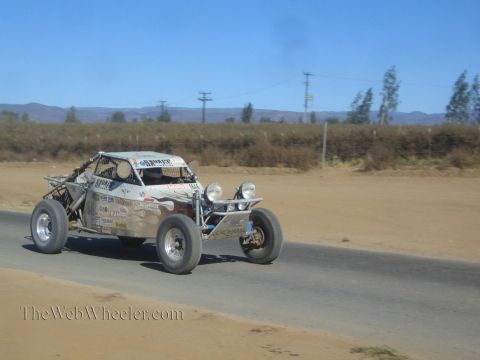1990 Ford Bronco - silver-gray off-road race buggy with twin round headlights speeds along a paved road in dry hill country.
