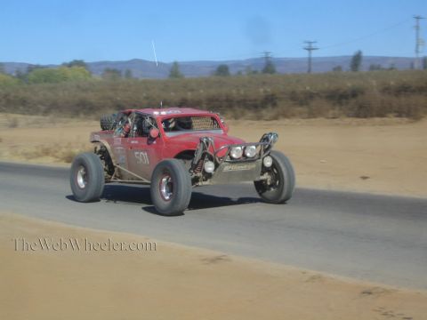 1990 Ford Bronco - dark red off-road race buggy numbered 501 with three roof lights speeds along a paved road kicking up dust.