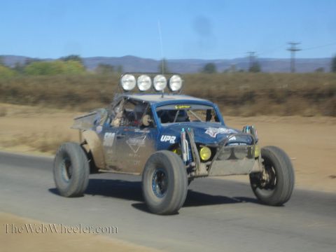 1990 Ford Bronco - blue and gray off-road race buggy numbered 500 with four roof lights races along a paved desert road.