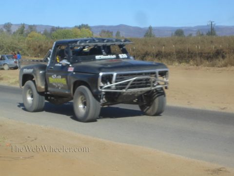 1990 Ford Bronco - black vintage Ford pickup built as an off-road race truck with tube bumper speeds along a paved desert road.